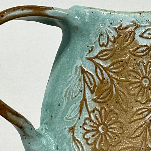 Close-up of a ceramic pitcher showing a light aqua glaze with a raised brown floral pattern and a brown handle, photographed against a plain light background.