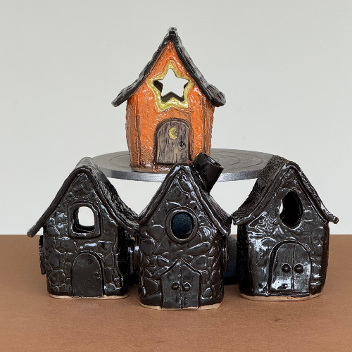 Three small decorative ceramic fairy houses with textured stucco-style surfaces, displayed on a turntable with an orange house on top. The houses are black with small colored accents on the doors and windows.